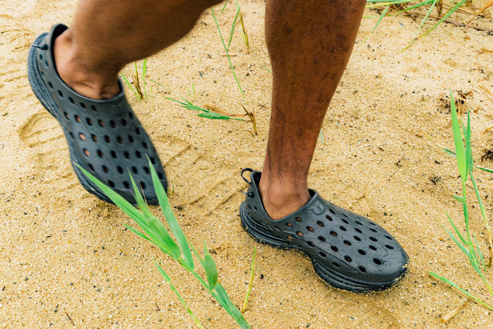 Person walking in black perforated shoes across sandy ground with sparse grass