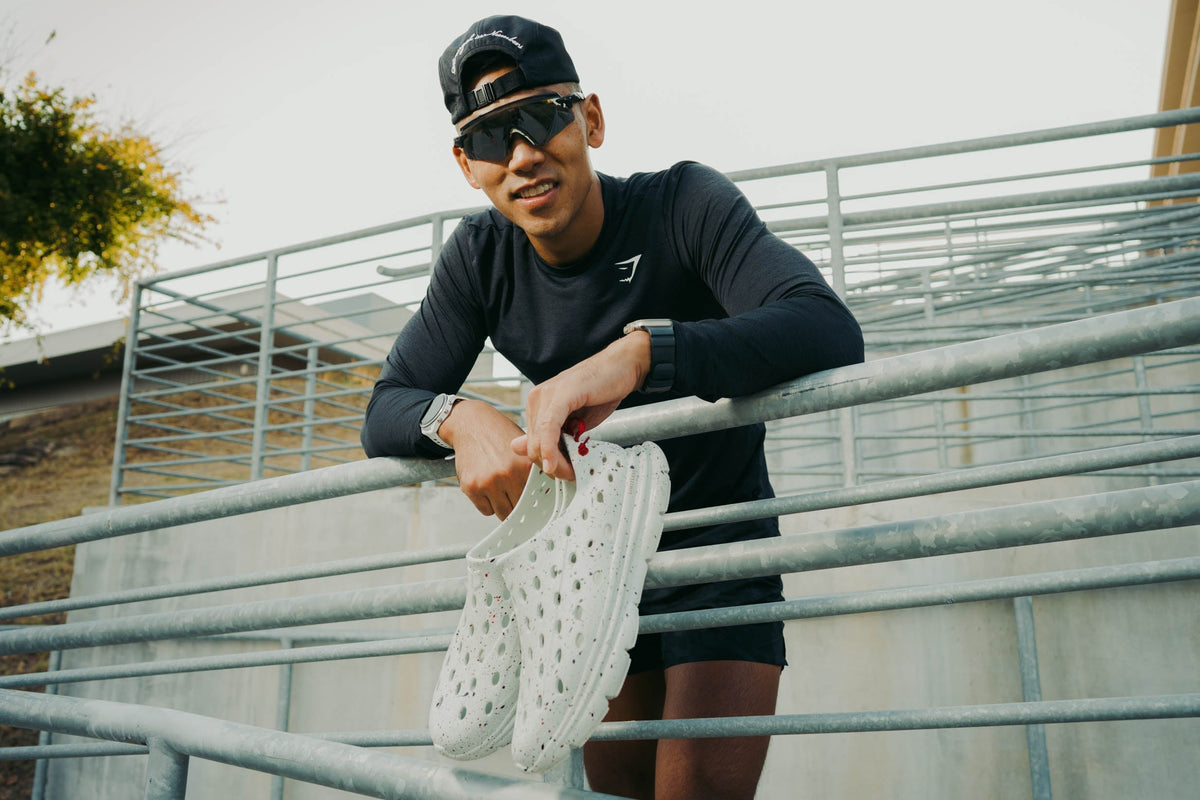 Man in black athletic clothing holding white athletic shoes beside outdoor railings