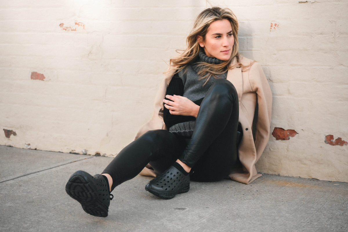 Woman sitting against a brick wall wearing black clogs, black leggings, and a beige coat