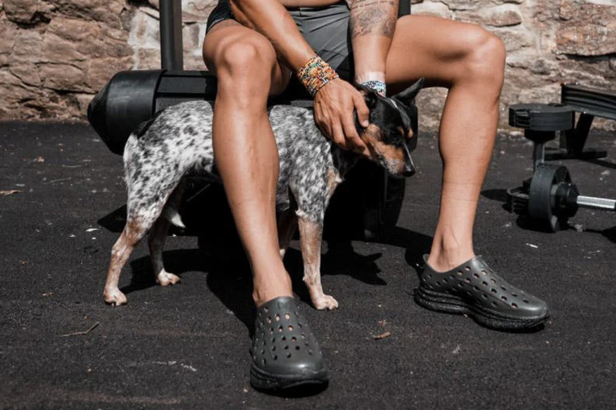 Person seated beside a dog wearing black clogs near outdoor exercise equipment