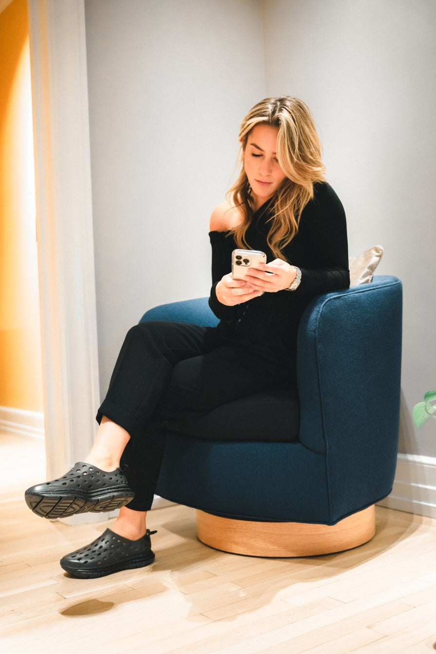 A woman sitting on a chair, checking her phone. Wearing Black Kane Revive work shoes.