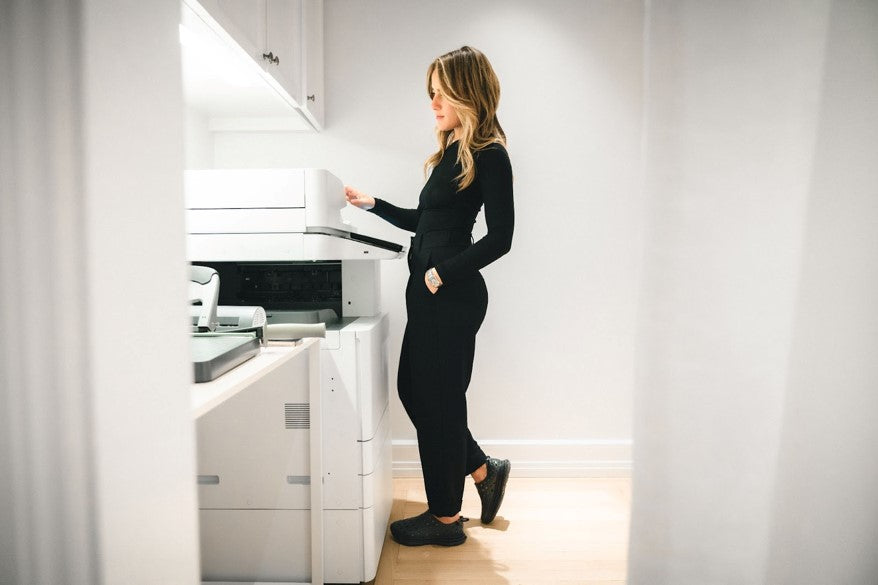 A teacher making photocopies by the printer, wearing stylish black Kane Revive recovery shoes.