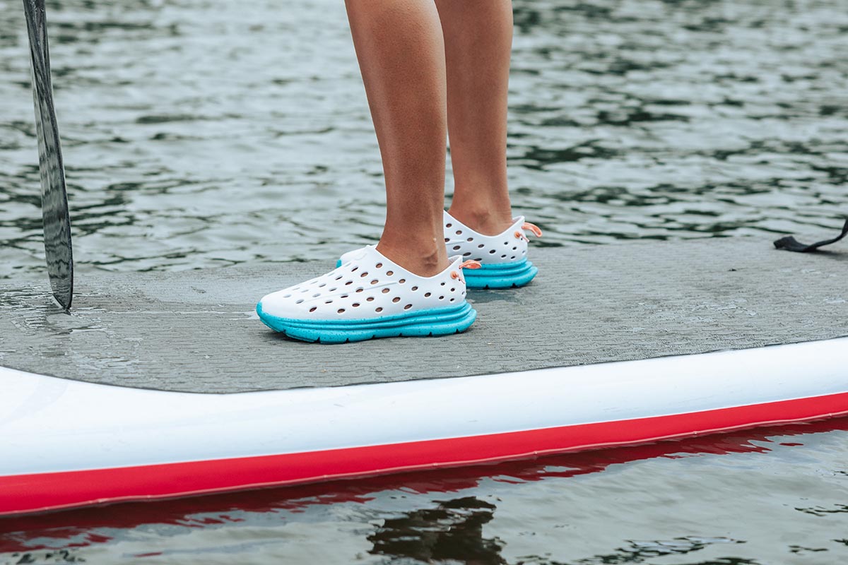 Person wearing white and turquoise clogs standing on a paddleboard over water