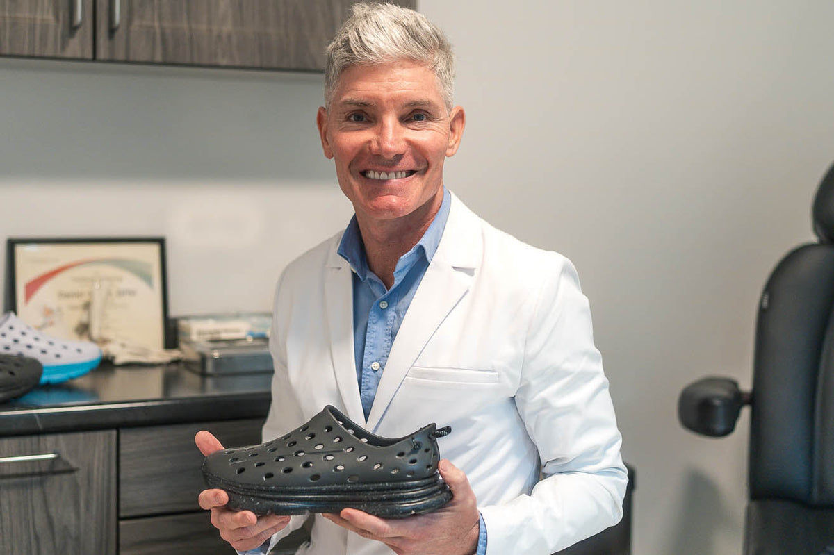 Smiling man in a white lab coat holds a black ventilated shoe in a clinical office