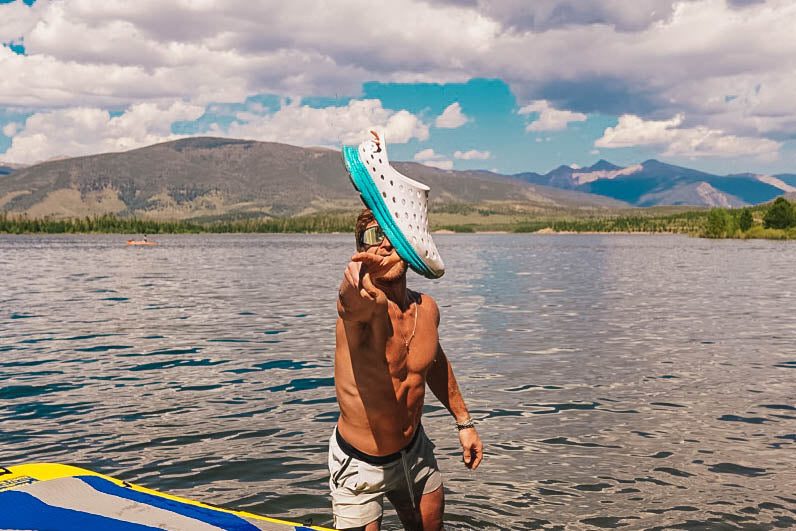 Shirtless man holds an oversized white and turquoise clog while standing in a mountain lake