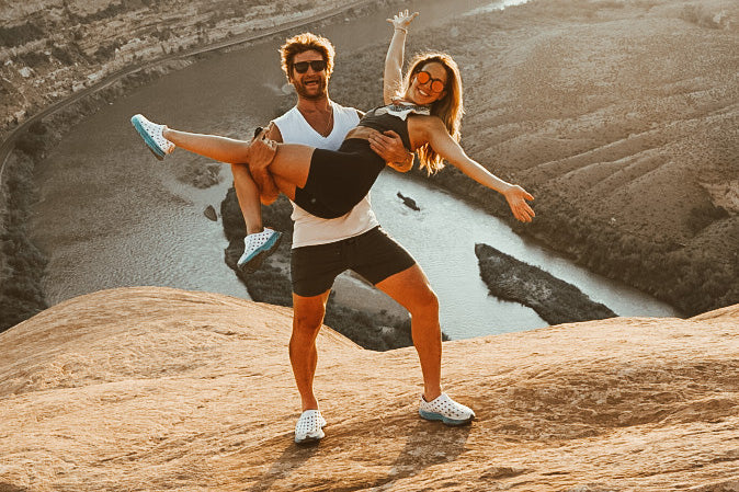 Couple playfully posing on a rocky overlook above a winding river, with one person carrying the other