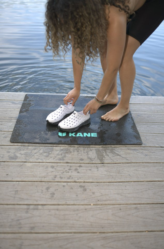 A woman putting on her white Kane revive shoes on top of a Kane Renew changing mat at the lake.
