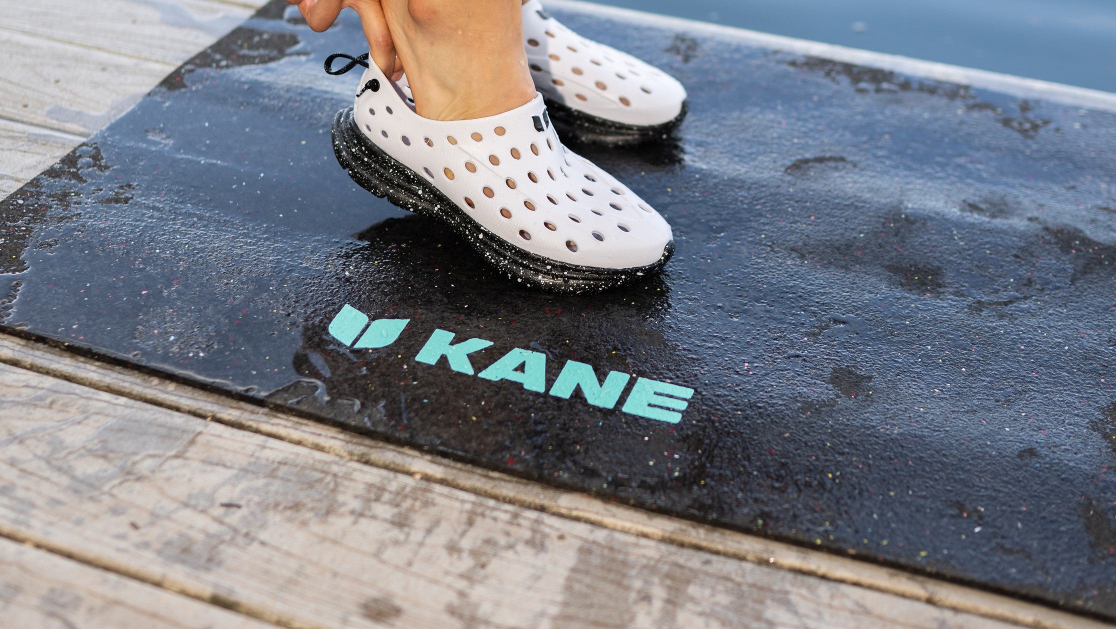 Slipping on Kane Revive shoes on top of a Kane Renew yoga mat. Placed on a wet deck to highlight the waterproof and non-slip features of Kane products.