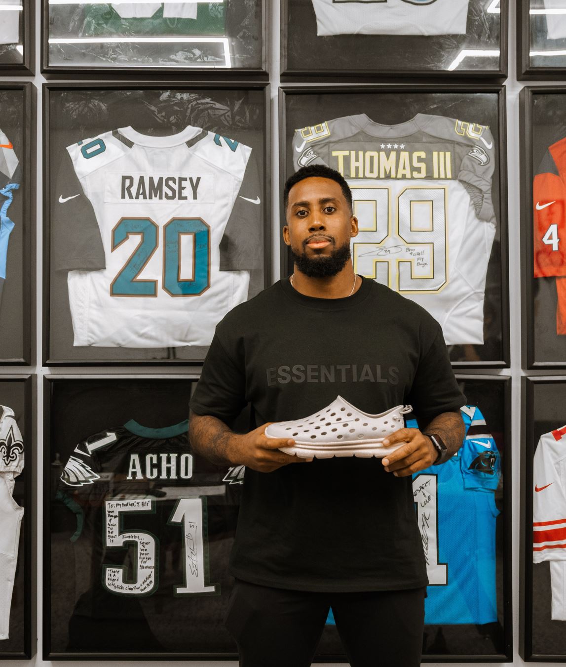 Portrait of Jeremy Hills posing with his Kane Revive recovery shoes. With a backdrop of collectable sports jerseys in cases behind him.