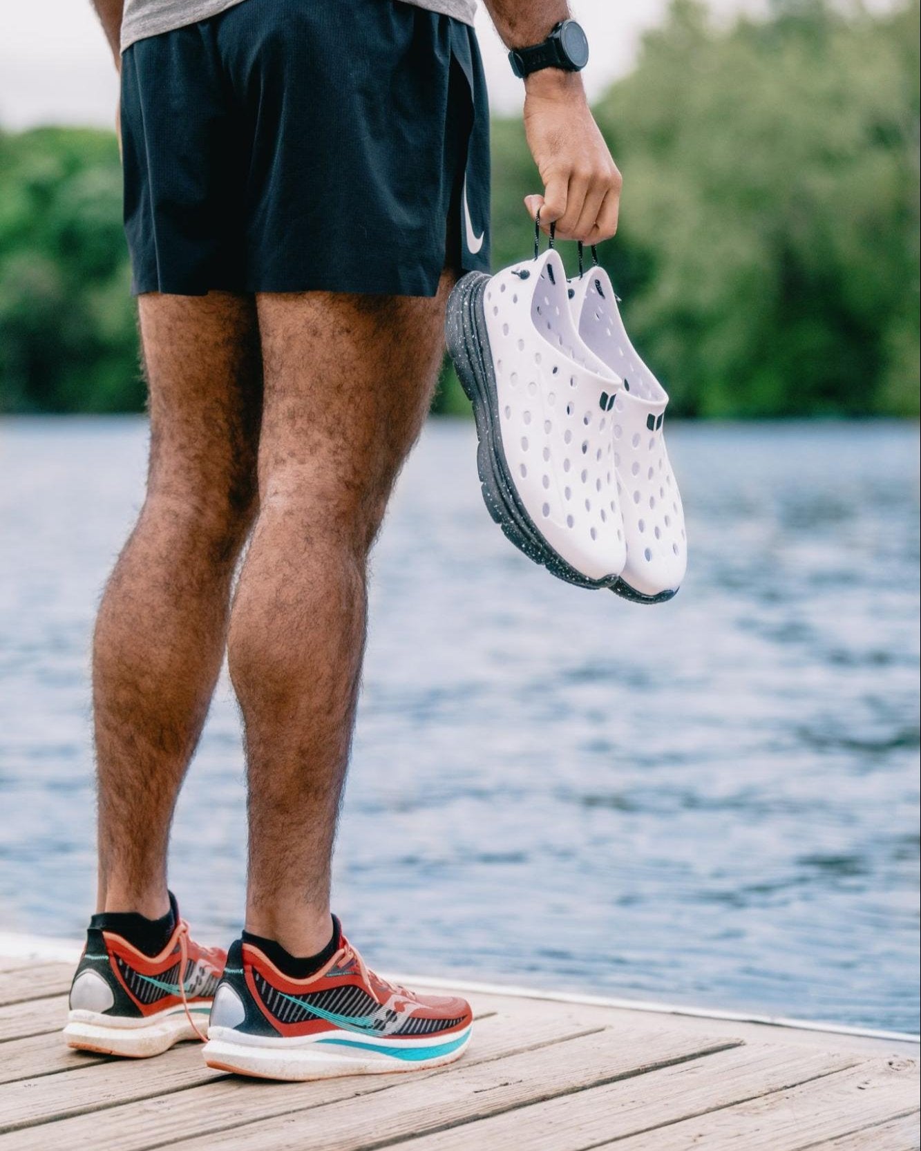 A man standing at the pier holding white Kane Revive shoes. He's looking out at the water.