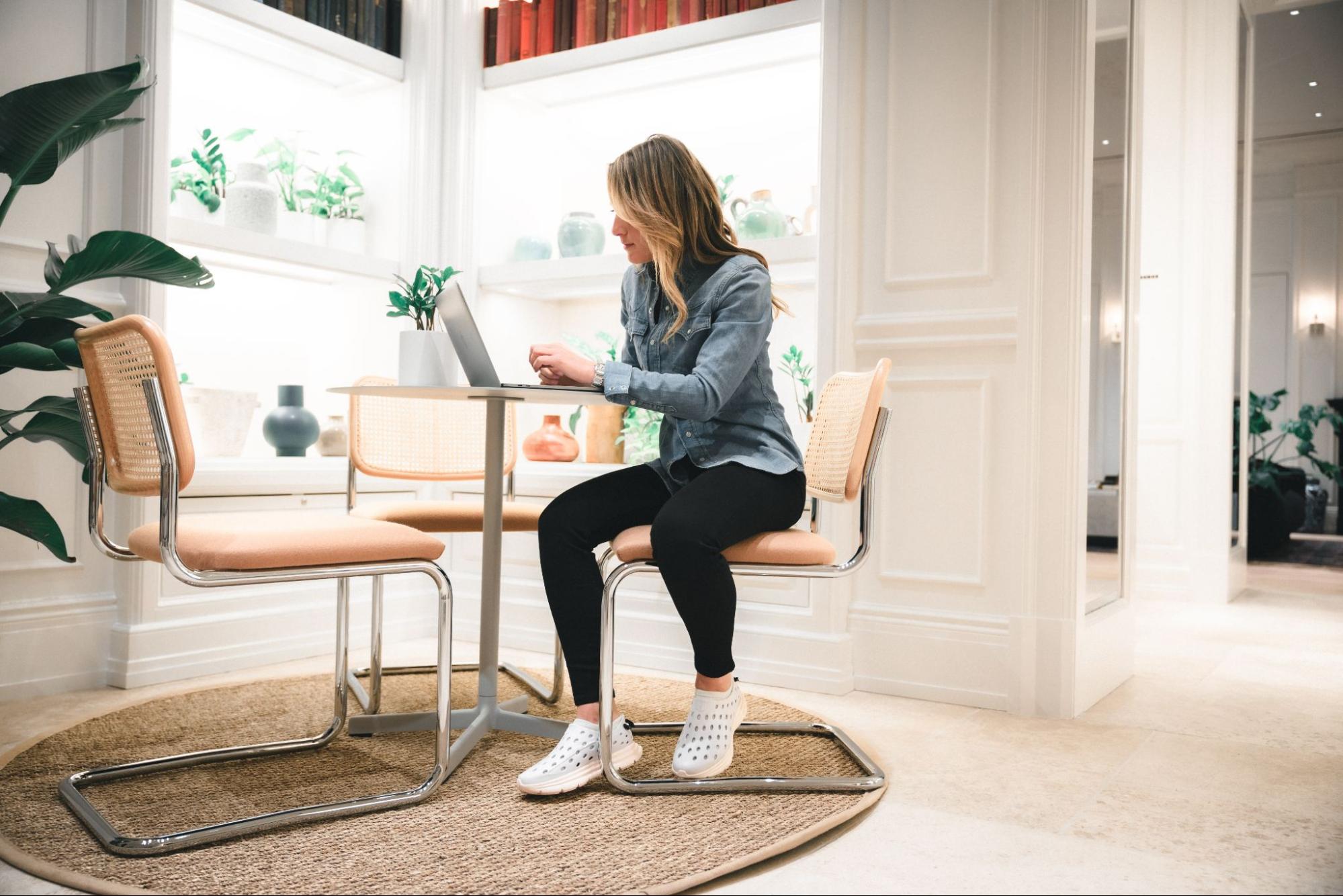 A woman sitting at a table working  on her laptop. She's wearing white Kane Revive recovery shoes.